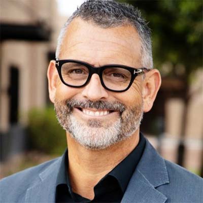 Man with salt & pepper hair and beard and black glasses smiles at the camera. He wears a gray suit and a black shirt.