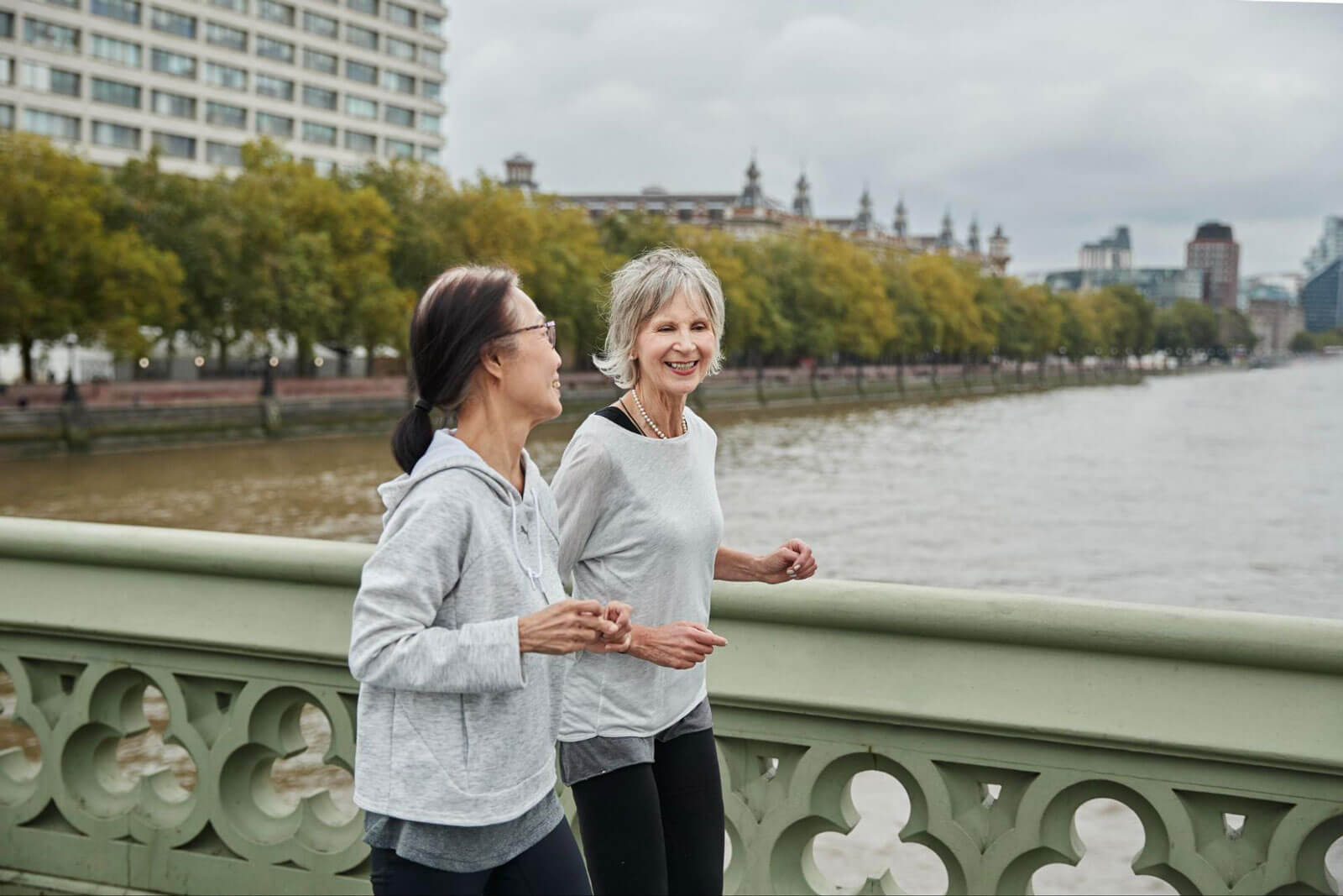Two older women in gray athletic wear smile and chat while jogging together along an ornate bridge railing with a river and city skyline visible in the background.
