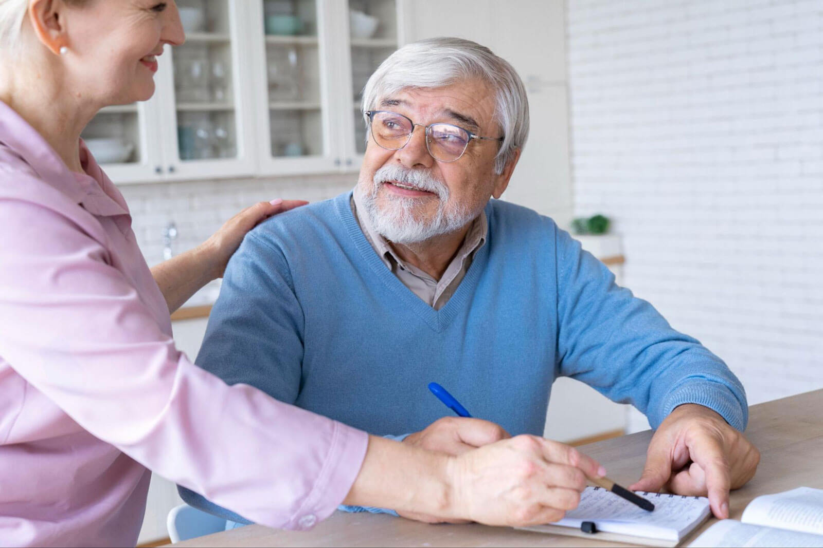 A woman rests her hand on an older man's shoulder while he writes in a notebook at a kitchen table.