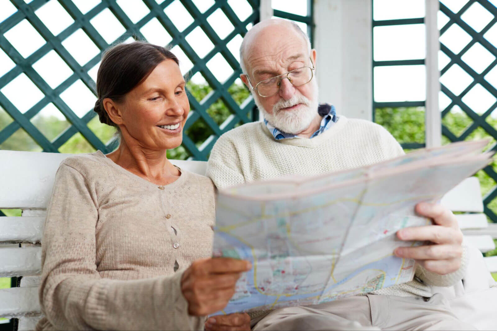 A smiling senior couple sits outdoors on a bench while reviewing a large map together.