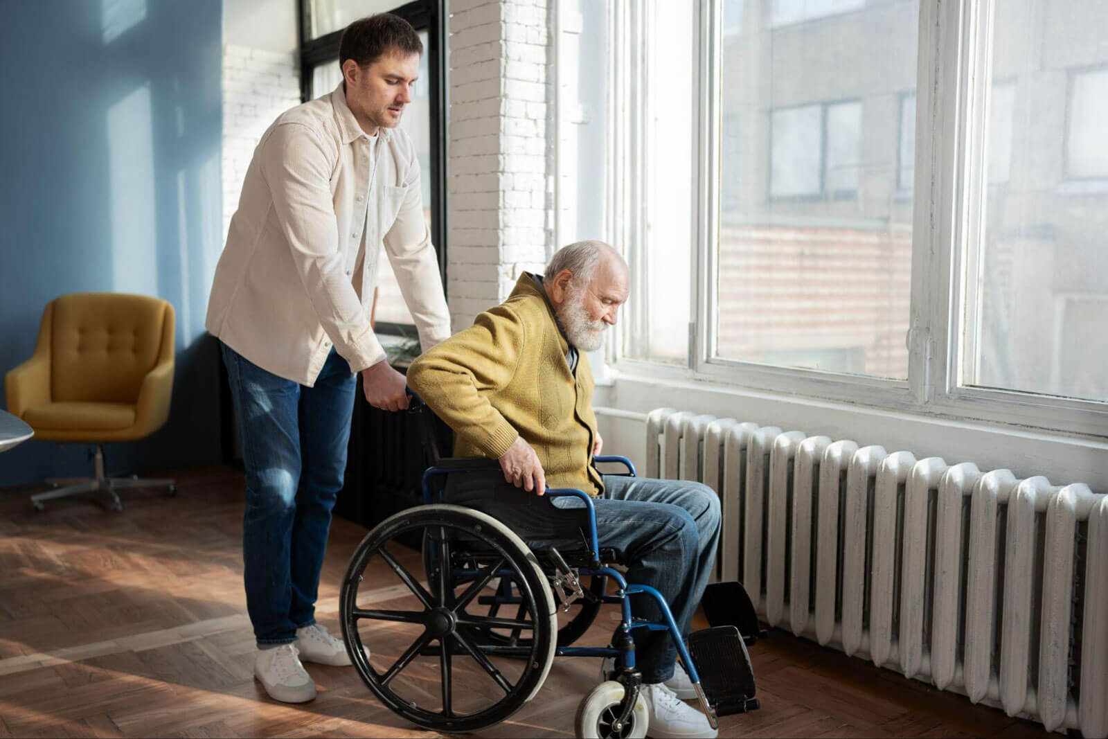 A man pushes an elderly man in a wheelchair past a window in a bright independent living facility.